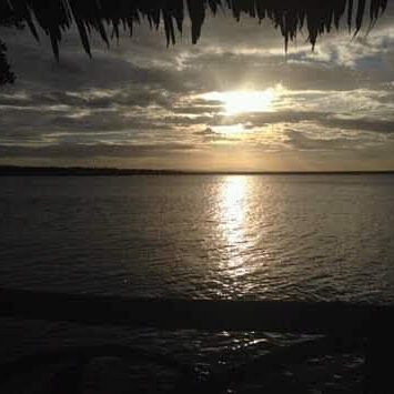 View of a golden sunset over a wide river or lake, seen from beneath a thatched roof, with a dark tree line on the horizon and light reflecting on the water
