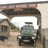 A safari jeep parked beneath the entrance gate of Tsavo East National Park, with a small building beside it