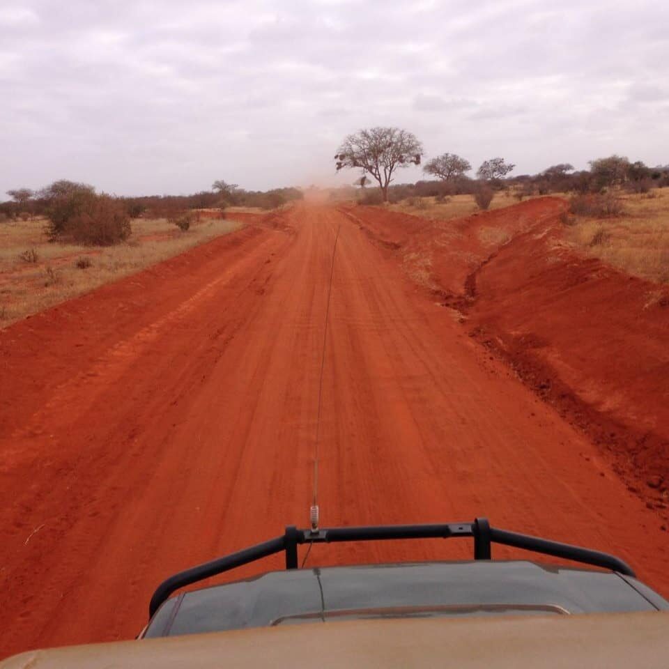 View from inside a safari vehicle driving along a red dirt road through dry savanna landscape under a cloudy sky.