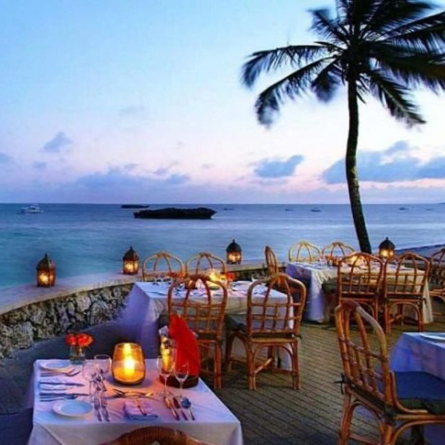 A romantic beachside restaurant with candlelit tables, wicker chairs, and a palm tree, overlooking the ocean at dusk