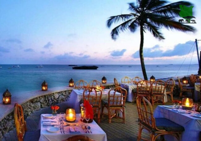 A romantic beachside restaurant with candlelit tables, wicker chairs, and a palm tree, overlooking the ocean at dusk