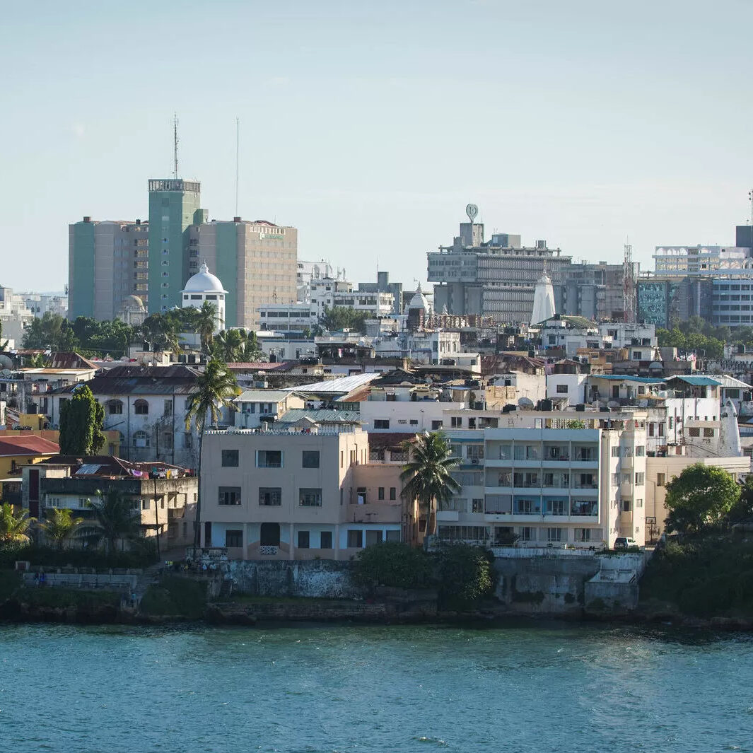 A coastal city skyline with a mix of older low-rise buildings along the waterfront and modern high-rise towers in the background, under a clear sky.