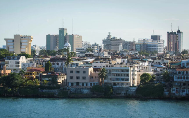 A coastal city skyline with a mix of older low-rise buildings along the waterfront and modern high-rise towers in the background, under a clear sky.