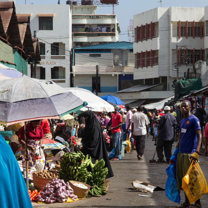 A busy street market in an East African town with vendors selling produce under umbrellas and pedestrians walking by