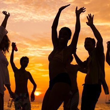 Silhouettes of friends dancing with raised arms on a beach at sunset, orange sky with a sailboat in the distance