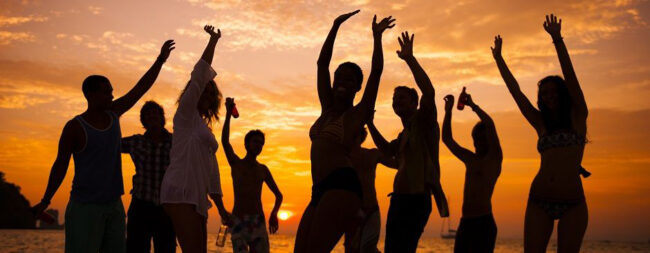Silhouettes of friends dancing with raised arms on a beach at sunset, orange sky with a sailboat in the distance