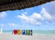 A colorful "I ❤ Diani" sign stands on the white sandy beach in front of turquoise ocean waters, with a thatched roof in the foreground and a sailboat visible on the horizon.