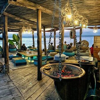 Rustic wooden beach lounge at dusk with hanging lights, low seating, and an ocean view