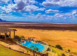 Aerial view of a kidney-shaped infinity pool with a stone-walled deck perched on a cliff edge, overlooking a vast golden savanna under a bright blue sky with scattered white clouds. A thatched-roof pavilion is visible in the left foreground